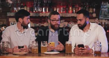 三个老朋友坐在啤酒酒吧里用智能手机。 有胡子的男人在他的智能手机上展示有趣的东西